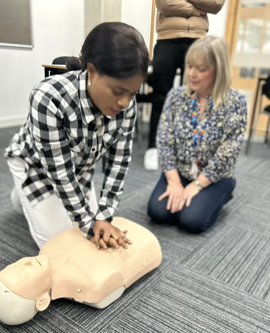 A woman in a black-and-white checkered shirt is administering CPR on a mannequin, while an instructor watches attentively beside her. A woman in a black-and-white checkered shirt is administering CPR on a mannequin, while an instructor watches attentively beside her.